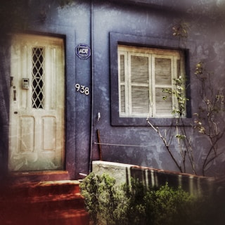 A weathered blue facade of a building features a white door with a diamond-patterned window and an adjacent number '938'. A security company sign is mounted near the door. Beside the door, there is a window with closed shutters. Sparse foliage and some bare branches can be seen in front of the building.