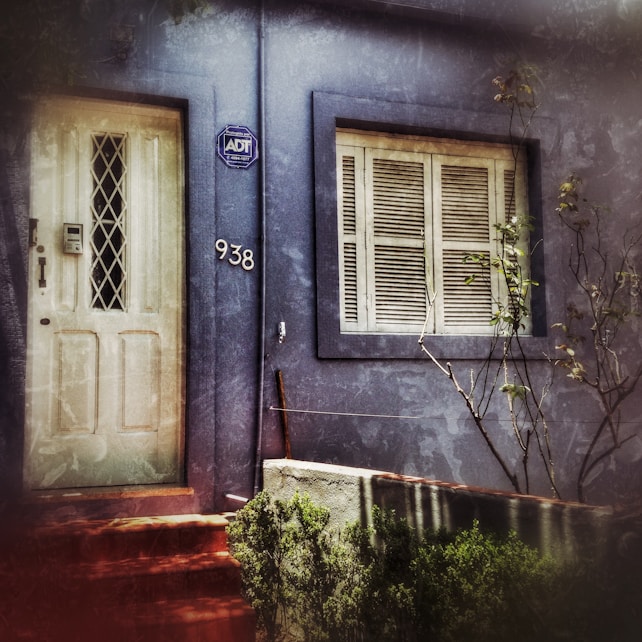 A weathered blue facade of a building features a white door with a diamond-patterned window and an adjacent number '938'. A security company sign is mounted near the door. Beside the door, there is a window with closed shutters. Sparse foliage and some bare branches can be seen in front of the building.