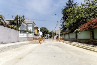 A quiet residential street lined with large trees and well-maintained houses. The road has some construction barriers and signage indicating possible road work. The sky is clear, suggesting a sunny day.