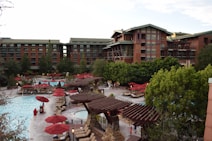An outdoor pool area surrounded by tall, rustic-style buildings. Numerous red umbrellas and lounge chairs are scattered around the pool, with several trees and greenery providing a natural backdrop. A few people are seen relaxing by the poolside.