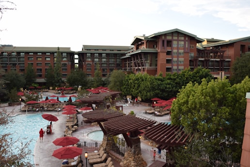An outdoor pool area surrounded by tall, rustic-style buildings. Numerous red umbrellas and lounge chairs are scattered around the pool, with several trees and greenery providing a natural backdrop. A few people are seen relaxing by the poolside.