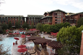An outdoor pool area surrounded by tall, rustic-style buildings. Numerous red umbrellas and lounge chairs are scattered around the pool, with several trees and greenery providing a natural backdrop. A few people are seen relaxing by the poolside.