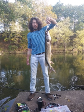 A person stands on a concrete platform by a calm body of water, holding a large fish. The background features lush green trees with leaves that have started to turn yellow, indicating early autumn. The person is dressed in a blue and white striped long-sleeve shirt and light blue jeans. Various fishing gear is scattered on the ground nearby.