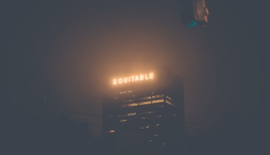 A tall building with bright, illuminated letters spelling 'EQUITABLE' on the top, shrouded in fog at night. The visibility of the surrounding area is low, and the glow from the signage creates a soft, atmospheric light against the dark background.