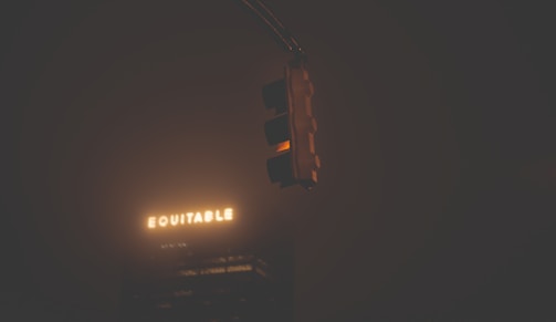 A dimly lit traffic light glows faintly against a dark background with a building showing the illuminated word 'EQUITABLE'. The scene is largely shrouded in gloom, giving an impression of nighttime or fog.