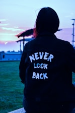 A person wearing a dark jacket with the words 'NEVER LOOK BACK' written on the back is sitting outdoors. The background includes a pergola, grass, and a colorful sky, likely during sunset or sunrise.