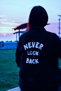 A person wearing a dark jacket with the words 'NEVER LOOK BACK' written on the back is sitting outdoors. The background includes a pergola, grass, and a colorful sky, likely during sunset or sunrise.
