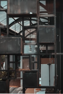 A modern interior space featuring a grid-like metal frame in the foreground. Through the frame, various elements such as a glass ceiling with geometric patterns, hanging light fixtures, and parts of the indoor decor are visible. The overall ambiance is industrial with an architectural focus.