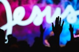 A silhouette of a person raising their hand in front of a vibrant background featuring the word 'Jesus' in large, flowing white script. The background displays a mixture of blue, pink, and purple hues.