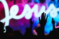 A silhouette of a person raising their hand in front of a vibrant background featuring the word 'Jesus' in large, flowing white script. The background displays a mixture of blue, pink, and purple hues.