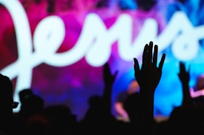 A silhouette of a person raising their hand in front of a vibrant background featuring the word 'Jesus' in large, flowing white script. The background displays a mixture of blue, pink, and purple hues.