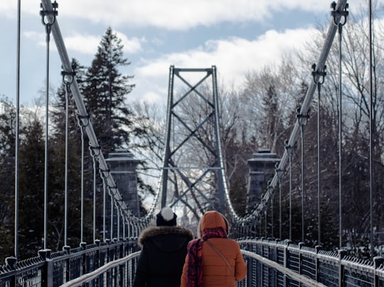 two persons walking on gray suspension bridge in between trees at daytime in Quebec Canada