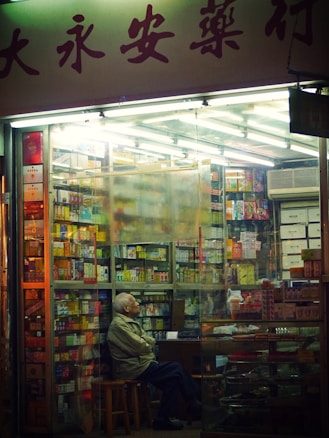 Inside a small, dimly lit shop filled with shelves of various products, an elderly man sits on a stool, appearing contemplative. The shop has fluorescent lighting, and the shelves are stocked with colorful boxes. The scene is captured through a glass window, outside of which Chinese characters are visible.