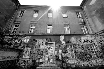 A weathered urban building adorned with various colorful graffiti covers a large portion of the facade. Sunlight beams down from above, creating a dramatic highlight on the structure. The walls are filled with vibrant street art, including tags, characters, and abstract designs, suggesting an expressive urban culture.