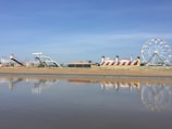 An amusement park is visible from a sandy beach with a calm body of water reflecting parts of the park. The scene includes a large Ferris wheel to the right, colorful slides, and a building with red and white stripes resembling a circus tent.