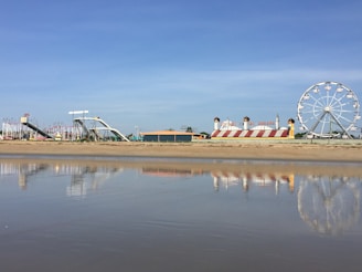 An amusement park is visible from a sandy beach with a calm body of water reflecting parts of the park. The scene includes a large Ferris wheel to the right, colorful slides, and a building with red and white stripes resembling a circus tent.