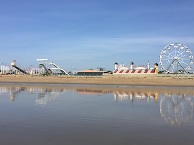 An amusement park is visible from a sandy beach with a calm body of water reflecting parts of the park. The scene includes a large Ferris wheel to the right, colorful slides, and a building with red and white stripes resembling a circus tent.