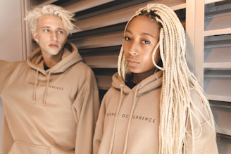Two individuals, one with light blonde hair and the other with long braided hair, are wearing matching beige hoodies with the text 'Change Occurrence.' They are standing against a metal louvered background.
