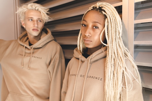 Two individuals, one with light blonde hair and the other with long braided hair, are wearing matching beige hoodies with the text 'Change Occurrence.' They are standing against a metal louvered background.