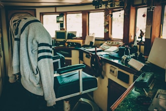 An interior view of a boat cockpit with a large steering wheel, various control panels, a chair draped with a sweatshirt, and windows providing natural light. The space is cluttered with electronic devices, books, and equipment scattered on countertops.