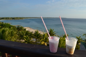Two plastic cups with long straws sit on a wooden railing, overlooking a tropical beach with clear blue skies and a calm sea. The shoreline curves gently into the distance, bordered by lush green palm trees. People can be seen enjoying the beach and swimming in the water.
