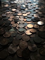 A large number of coins scattered across a surface, creating a textured and uneven landscape. The coins vary slightly in color and appear mostly metallic with shadows and reflections enhancing their details.