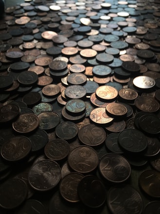 A large number of coins scattered across a surface, creating a textured and uneven landscape. The coins vary slightly in color and appear mostly metallic with shadows and reflections enhancing their details.