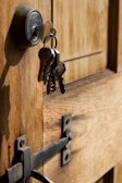 A close-up view of a wooden door with a metal lock and keys hanging from it. The keys are inserted into the lock, and the metal latch on the door is visible.