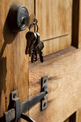 A close-up view of a wooden door with a metal lock and keys hanging from it. The keys are inserted into the lock, and the metal latch on the door is visible.