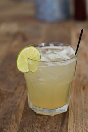 A refreshing drink served in a glass with ice cubes and a slice of lime on the rim, placed on a wooden surface. A black straw is also in the glass, suggesting the drink is ready to be enjoyed.