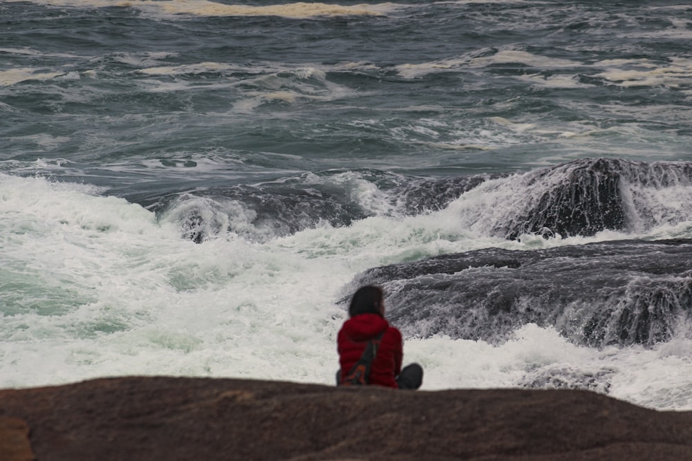 photo of person sitting on rock watching sea