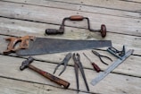 A collection of vintage hand tools lies on a wooden surface. The assortment includes a rusty handsaw, a hand drill with a wooden handle, pliers, a hammer with a wooden handle, a flat-head screwdriver, a combination square, and other metal tools. The tools have an aged appearance, indicating frequent use over the years.