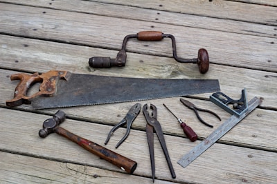 A collection of vintage hand tools lies on a wooden surface. The assortment includes a rusty handsaw, a hand drill with a wooden handle, pliers, a hammer with a wooden handle, a flat-head screwdriver, a combination square, and other metal tools. The tools have an aged appearance, indicating frequent use over the years.