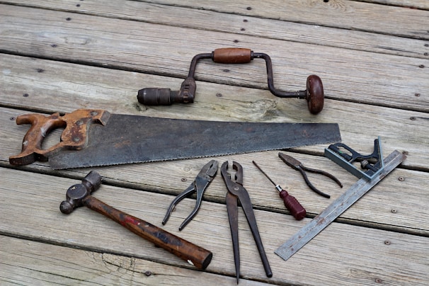 A collection of vintage hand tools lies on a wooden surface. The assortment includes a rusty handsaw, a hand drill with a wooden handle, pliers, a hammer with a wooden handle, a flat-head screwdriver, a combination square, and other metal tools. The tools have an aged appearance, indicating frequent use over the years.