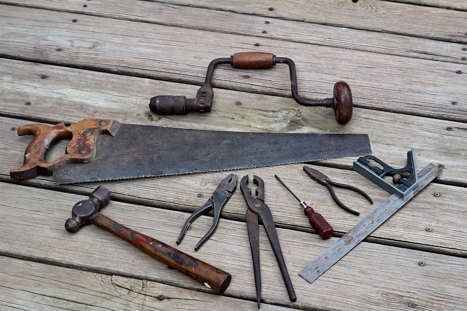 A collection of vintage hand tools lies on a wooden surface. The assortment includes a rusty handsaw, a hand drill with a wooden handle, pliers, a hammer with a wooden handle, a flat-head screwdriver, a combination square, and other metal tools. The tools have an aged appearance, indicating frequent use over the years.