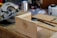 A wooden workbench in a workshop with various tools and a piece of wood standing upright on the table. A circular saw is positioned on the left, and there are additional tools like a metal ruler and a container in the background.