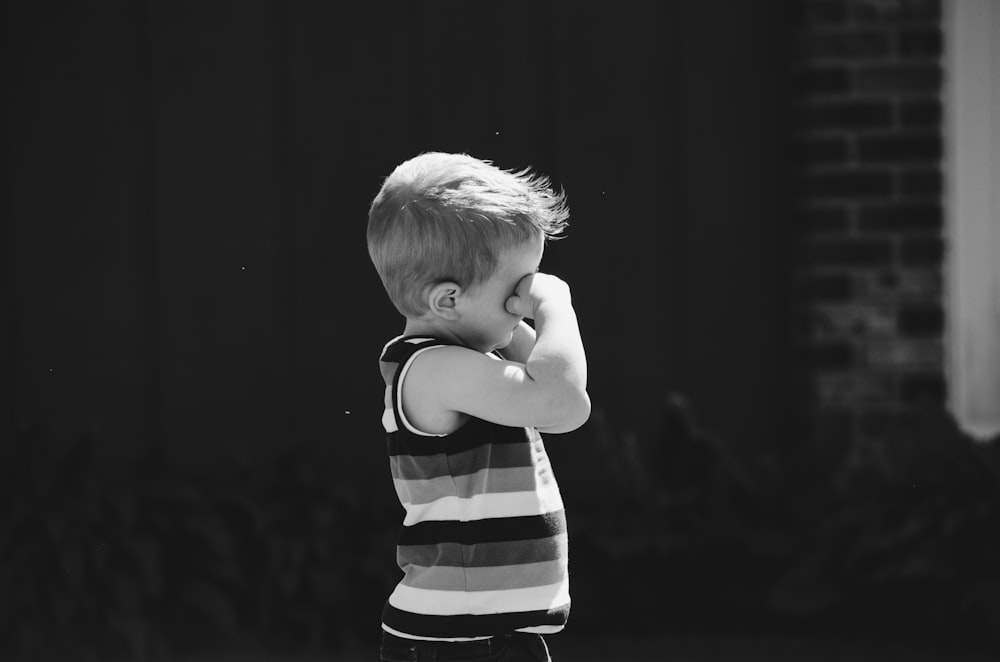 grayscale photo of boy covering of eye while standing behind concrete wall