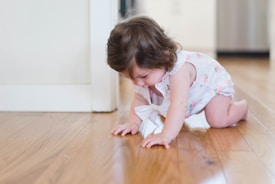 A baby in a floral-patterned onesie is crawling on a wooden floor, appearing focused and curious about the surface. The background is softly blurred, suggesting a home setting.