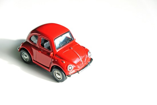 A miniature red vintage car model displayed on a clean, white background. The car has a shiny finish, detailed windows, and black tires. Its design is reminiscent of classic cars.