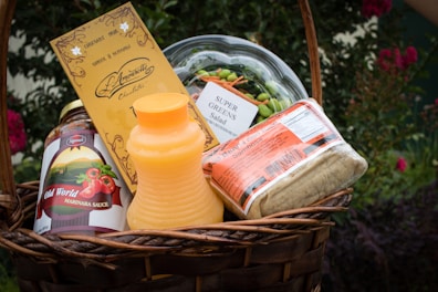 A woven basket filled with various food items including a bottle of marinara sauce, a carton of orange juice, a package of wraps, a container of super greens salad, and a bar of chocolate.