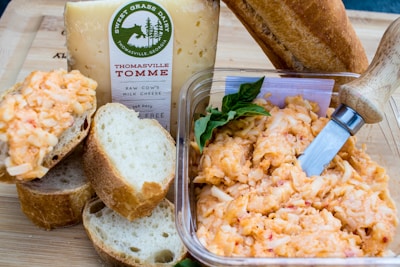 A wooden cutting board displays a large block of Sweet Grass Dairy cheese, labeled Thomasville Tomme. Beside it, there's a small loaf of crusty bread and several slices. A clear plastic container holds a cheesy spread with visible shreds, accompanied by a wooden-handled cheese knife. Fresh green basil leaves are tucked near the spread.
