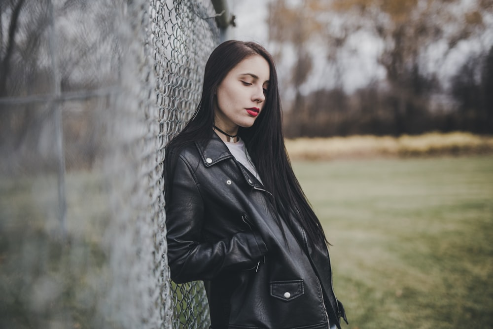 woman leaning on cyclone fence with her right hand on her right jacket pocket