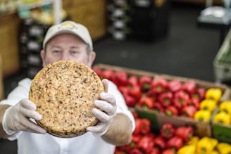A person wearing a cap and gloves holds up a round cheese with visible herbs or spices embedded in it. In the background, there are colorful red and yellow bell peppers neatly stacked in boxes.