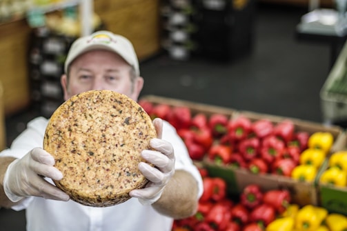 A person wearing a cap and gloves holds up a round cheese with visible herbs or spices embedded in it. In the background, there are colorful red and yellow bell peppers neatly stacked in boxes.