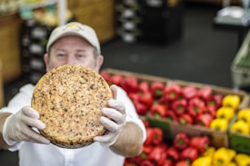 A person wearing a cap and gloves holds up a round cheese with visible herbs or spices embedded in it. In the background, there are colorful red and yellow bell peppers neatly stacked in boxes.