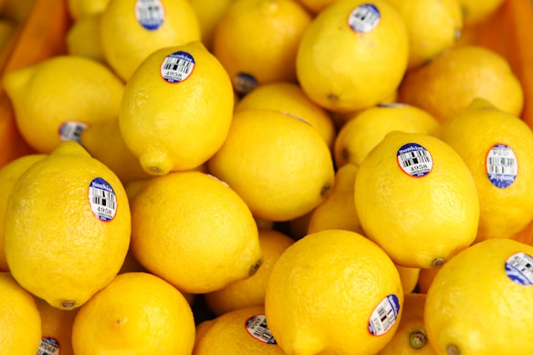 A pile of bright yellow lemons with blue stickers displaying pricing and product information. They are closely packed together in a market setting.