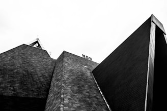 A black and white photograph showcasing a modern architectural structure with angular, geometric shapes and textured brick surfaces. The sharp lines and contrasting tones create a striking and bold visual effect against the pale sky.
