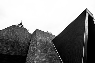A black and white photograph showcasing a modern architectural structure with angular, geometric shapes and textured brick surfaces. The sharp lines and contrasting tones create a striking and bold visual effect against the pale sky.