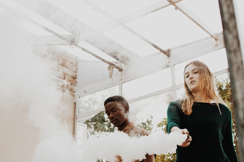Two people stand beneath a glass ceilinged structure, surrounded by billows of white smoke. The setting appears to be open and airy, with natural light filtering through the roof. One individual holds a device emitting the smoke.