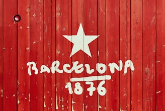 Wooden planks painted red form a background for the word 'Barcelona' with the number '1876' written underneath in white paint. A white star is displayed above the text, giving the composition a vintage or rustic look.